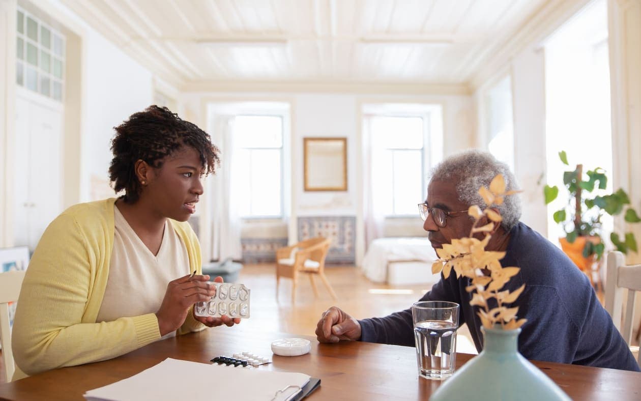 A young woman and an older man at the dining table at home, going over his tablets for the day with a glass of water