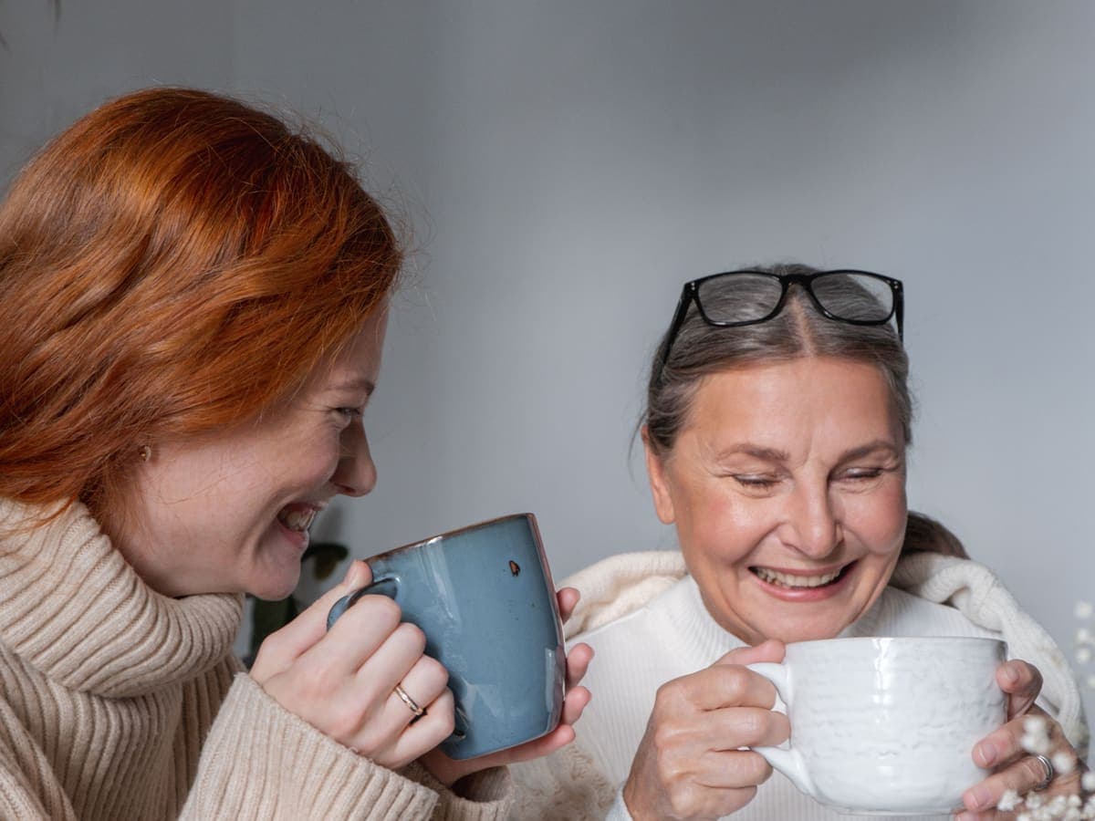A woman and her mother laughing together over two mugs of tea
