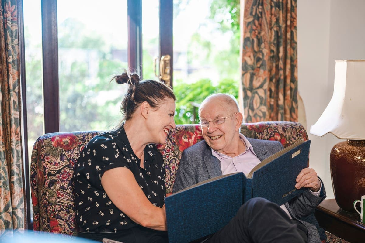 A woman and an older man laughing together over a photo album on a sofa