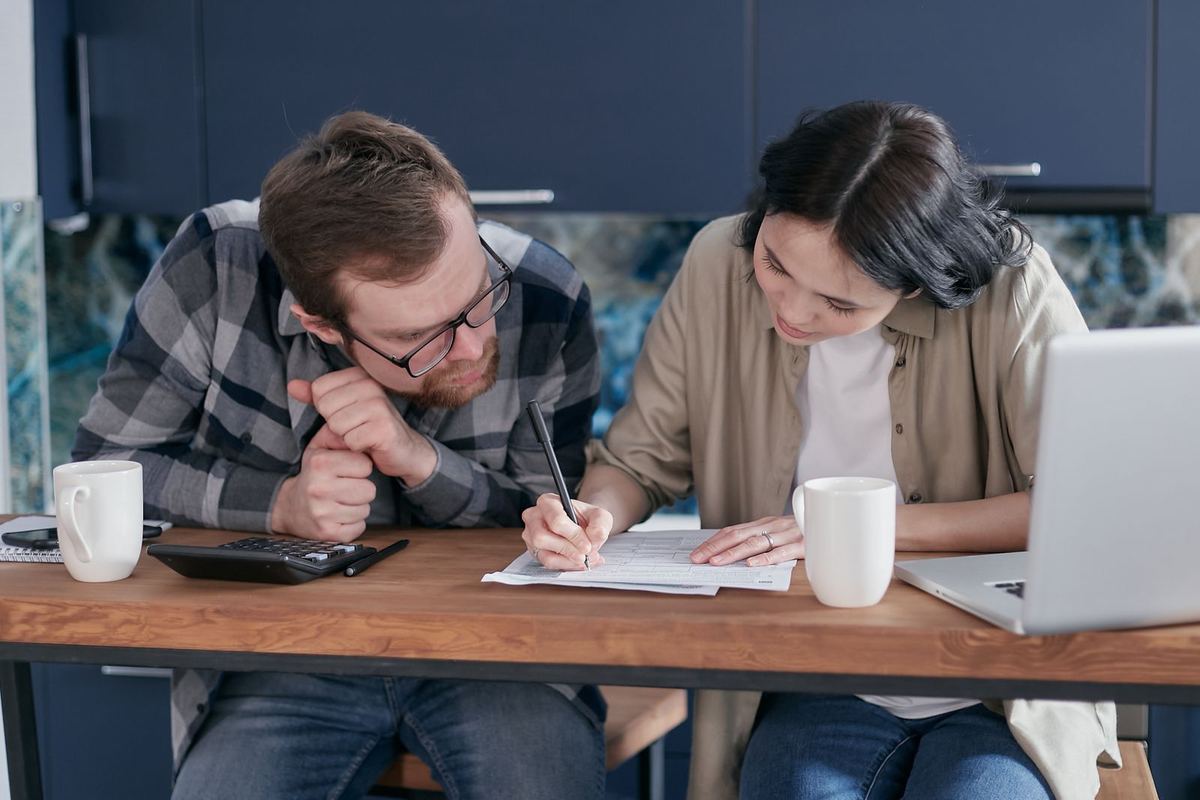Two adults at a kitchen table going through paperwork together, with coffee and a laptop.
